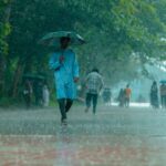A man in blue walking under heavy rain with umbrella on a tree-lined path.